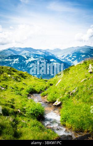 Un ruisseau de montagne limpide coule gracieusement à travers des pentes verdoyantes et herbeuses, menant le regard vers de majestueux sommets lointains. Banque D'Images