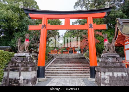 Les visiteurs montent des marches à travers une porte torii orange vibrante, gardée par des renards Inari en pierre, vers un sanctuaire shinto japonais. Banque D'Images