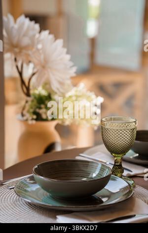 Superbe décor de table avec un verre à vin teinté vert, un bol gris sur des assiettes et un vase orné de fleurs blanches dans un cadre moderne de salle à manger Banque D'Images