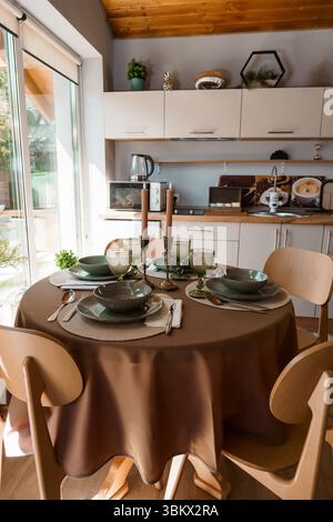 Une table ronde avec vaisselle verte élégante, draps beiges chauds et chaises en bois dans une cuisine moderne et lumineuse Banque D'Images
