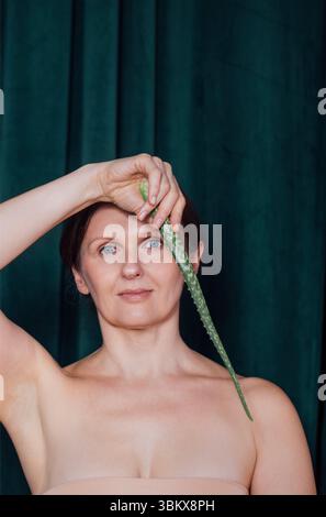 Une femme d'âge moyen tient une longue feuille d'aloès verticalement à côté de son visage, exprimant calme et harmonie. Une jeune femme avec une feuille verte pose contre un d. Banque D'Images