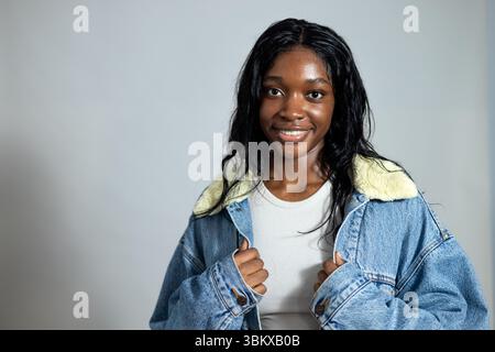 Portrait de studio à la taille haute d'une jeune femme adulte souriant à la caméra. Elle porte des vêtements décontractés, debout sur un fond de studio neutre. Banque D'Images