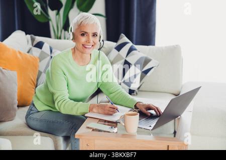 Femme mature souriante portant un pull vert avec casque travaillant à la maison sur un ordinateur portable, assis sur un canapé dans le confort Banque D'Images