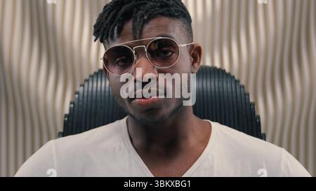 Portrait African American Guy homme ethnique masculin mettant sur des lunettes de soleil lunettes portant regarder la caméra assis dans futuriste blanc studio futurisme Banque D'Images