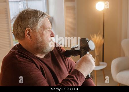 Homme âgé utilisant un pistolet de massage portatif électrique pour soulager la douleur du bras thérapie de récupération musculaire et soutien de bien-être à la maison Banque D'Images