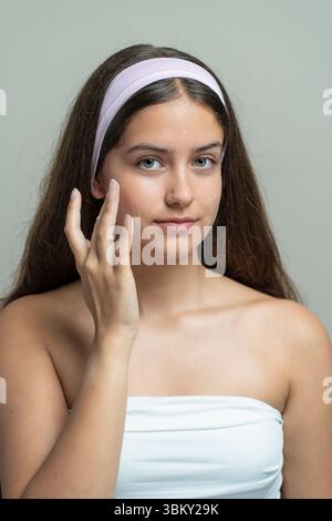 Une belle jeune femme avec un bandeau regarde l'appareil photo pendant qu'elle applique un hydratant ou une crème pour le visage. Concept d'une routine quotidienne de soin de la peau Banque D'Images