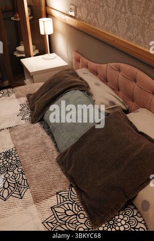 Scène de chambre confortable avec un lit soigneusement fait, des oreillers décoratifs et une lampe de chevet brillante chaleureusement. Un papier peint élégant et des textures douces créent un confort Banque D'Images