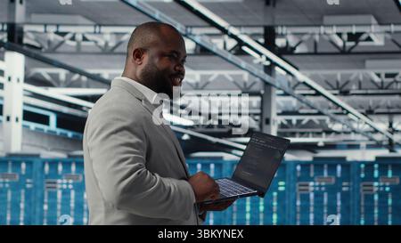 Un administrateur système de centre de données joyeux révise le code des scripts d'automatisation. Employé de la salle des serveurs Jolly exécutant des diagnostics sur l'équipement, effectuant la détection prédictive des erreurs, caméra B. Banque D'Images