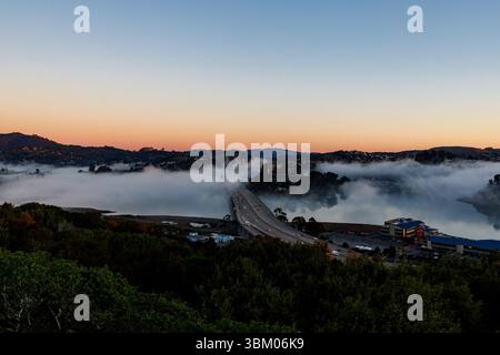 Une vue aérienne montre des voitures traversant un pont au-dessus d'une étendue d'eau, avec du brouillard qui roule autour des collines et des bâtiments. Banque D'Images