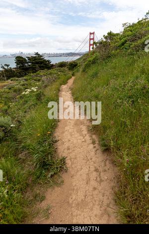 Un chemin de terre serpente à travers une colline herbeuse, menant au Golden Gate Bridge et à la skyline de San Francisco au loin, offrant une vue panoramique pour les randonneurs et les passionnés de nature. Banque D'Images