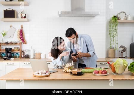Jeune couple regardant un smartphone ensemble tout en mangeant et en travaillant dans une cuisine moderne. Ordinateur portable et nourriture saine sur le comptoir. Partager un moment heureux Banque D'Images