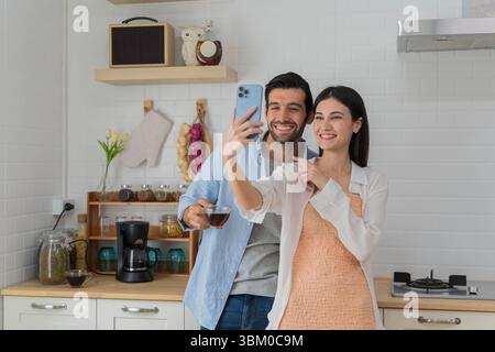 Jeune couple prenant selfie dans la cuisine confortable tout en tenant le café. À la fois souriant et profitant d'une matinée amusante et détendue ensemble. Capturer des souvenirs heureux, Banque D'Images