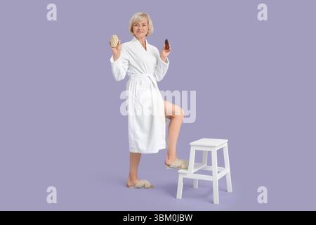 Belle femme heureuse mature en peignoir avec brosse de massage et bouteille de produit cosmétique sur chaise sur fond violet Banque D'Images