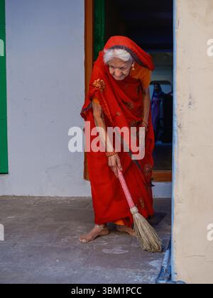 Inde, Delhi, 25 juin 2025. Femme âgée en sari rouge balayant le sol devant sa maison à Paharganj. Banque D'Images