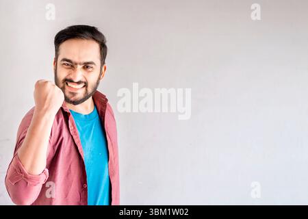 L'image montre un jeune homme avec une barbe, portant une chemise marron sur un t-shirt bleu. Il exprime la victoire ou l'excitation avec un poing levé, showin Banque D'Images