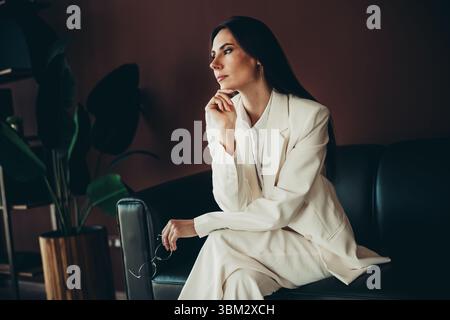 Femme d'affaires élégante portant un costume blanc, assise sur un canapé, contemplant avec une expression réfléchie dans un cadre de bureau élégant Banque D'Images