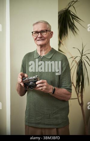 Homme âgé avec des lunettes souriant tout en tenant un appareil photo vintage, debout à l'intérieur devant une plante en pot, portant une chemise verte et regardant le contenu Banque D'Images