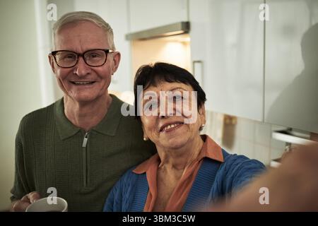 Portrait de couple âgé souriant tout en posant pour selfie dans une cuisine lumineuse. Homme portant des lunettes tenant la tasse à café et femme dans le cardigan coloré appréciant le moment Banque D'Images