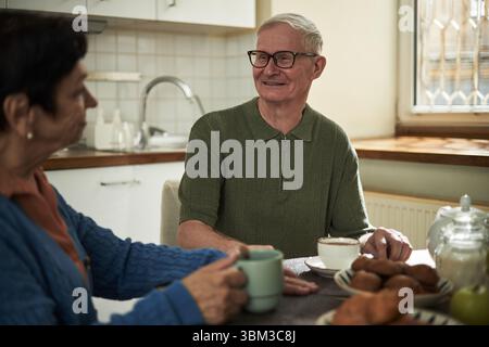 Homme senior souriant tout en prenant le petit déjeuner avec sa femme dans le cadre confortable de la cuisine avec lumière naturelle chaude coulant à travers la fenêtre Banque D'Images