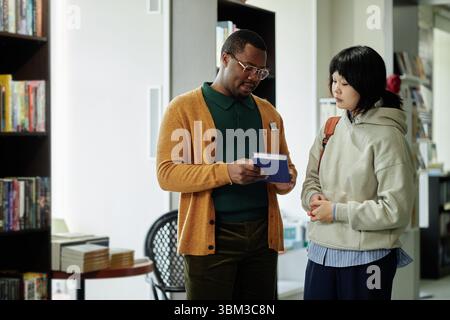 Homme noir d'âge moyen aidant jeune adulte asiatique femme choisir livre Banque D'Images