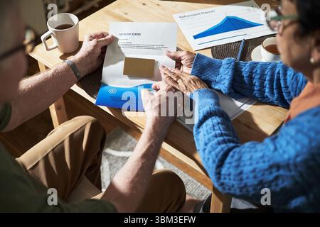 Couple senior ouvrant enveloppe avec carte de crédit, tout en examinant les plans de retraite à la maison, avec table et documents en arrière-plan, créant un moment de prise de décision partagée Banque D'Images