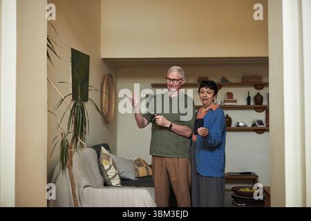 Couple de personnes âgées tenant des dispositifs tout en se tenant debout dans un salon confortable et bien décoré. Paire souriante engageant avec la technologie, entourée de plantes, d'étagères et de sièges confortables Banque D'Images