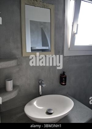 Intérieur minimaliste de la salle de bains avec mur gris clair, lavabo blanc et miroir. La lumière naturelle pénètre par une petite fenêtre. Banque D'Images