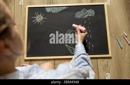 fille dessine la fleur et le soleil avec la craie colorée sur le tableau noir. dessin simple pour enfants sur tableau de craie noire Banque D'Images