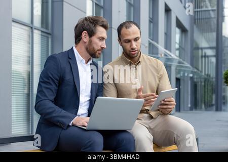 Deux hommes d'affaires divers en costumes discutent des affaires sur un banc à l'extérieur d'un immeuble de bureaux moderne, en utilisant un ordinateur portable et une tablette. Banque D'Images