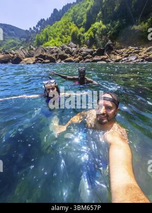 Trois amis portant un équipement de plongée en apnée et profitant de l'eau rafraîchissante de l'océan près d'une côte rocheuse Banque D'Images