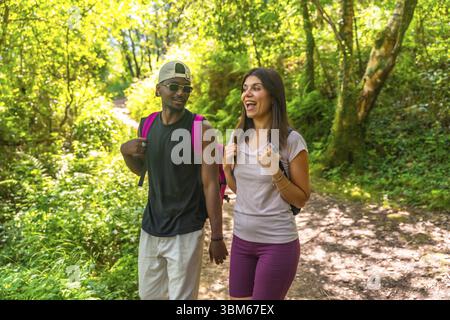 Couple interracial de randonneurs appréciant une conversation tout en marchant sur un sentier dans une forêt verdoyante Banque D'Images