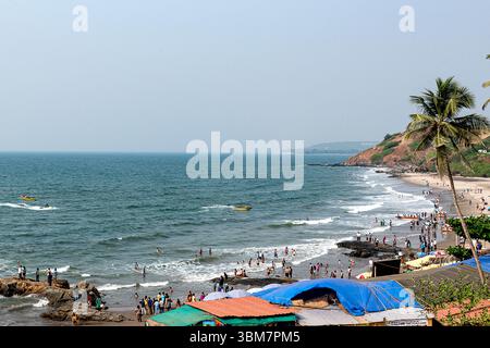 À Anjuna Beach à Goa, en Inde, de douces vagues se battent contre des formations rocheuses accidentées le long de la côte tandis que de nombreux visiteurs s'engagent dans des activités en bord de mer Banque D'Images
