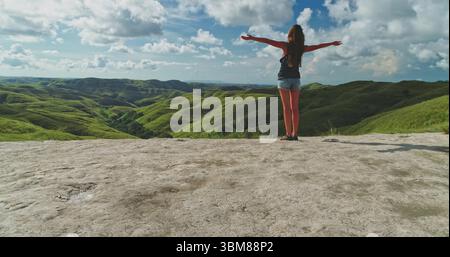 Jeune femme debout, les bras tendus, embrassant la beauté des collines verdoyantes et le vaste paysage de l'île de Sumba, une destination touristique populaire en Indonésie Banque D'Images
