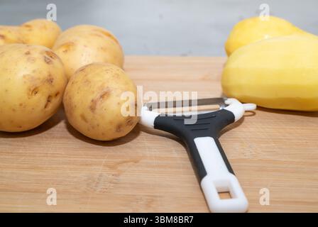 Une vue rapprochée montrant des pommes de terre pelées et non pelées aux côtés d'un éplucheur sur une planche à découper rustique en bois Banque D'Images