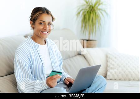Femme brésilienne positive dans des vêtements décontractés assis sur un canapé à la maison, souriant tout en tenant une carte de crédit et en utilisant un ordinateur portable. Elle aime les achats en ligne dans un cadre détendu et confortable dans un salon Banque D'Images