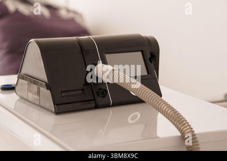 Gros plan d'un appareil de PPC noir avec tuyau attaché reposant sur une table de nuit blanche, représentant le traitement de l'apnée du sommeil, la technologie médicale au chevet du patient, et Banque D'Images