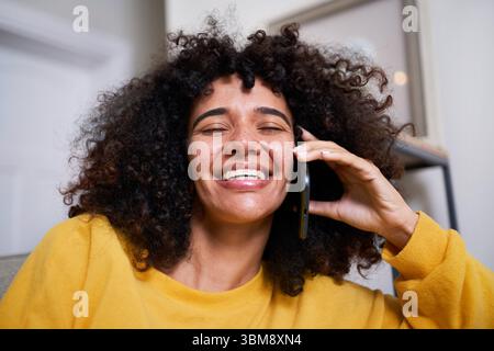 Femme joyeuse dans un pull jaune souriant tout en ayant une conversation téléphonique à la maison Banque D'Images