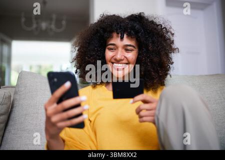 Heureuse jeune femme assise sur un canapé faisant un achat en ligne avec smartphone et carte de crédit Banque D'Images