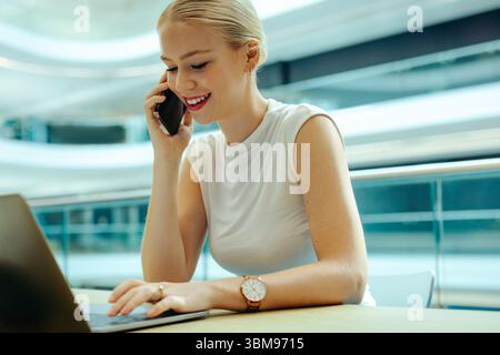 Une femme d'affaires blonde confiante parle sur son téléphone tout en travaillant sur son ordinateur portable dans un environnement de bureau moderne. Sa tenue professionnelle et ses expres concentrés Banque D'Images