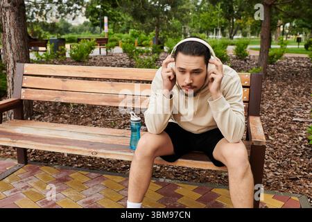 Un jeune homme en sweat à capuche est assis sur un banc, écoutant de la musique après l'entraînement en plein air au milieu de la verdure. Banque D'Images
