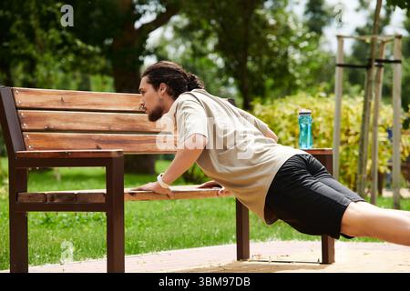 Un jeune homme en forme avec une barbe aime faire de l'exercice à l'extérieur en effectuant des pompes sur un banc. Banque D'Images