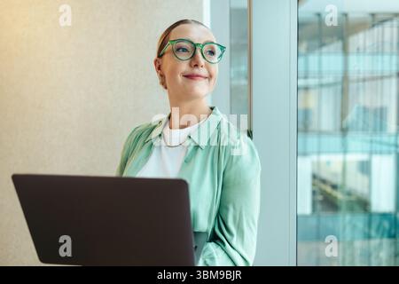 Une jeune femme professionnelle avec des lunettes tient en toute confiance un ordinateur portable. Elle dégage un sentiment de motivation et de disponibilité, appuyée contre un verre w Banque D'Images