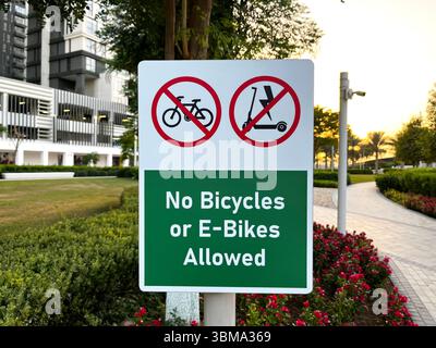 Panneau de parc urbain interdisant les vélos et les vélos électriques, avec des fleurs, une passerelle et des bâtiments modernes au coucher du soleil. Banque D'Images