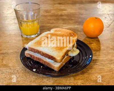 Petit-déjeuner de base servi dans une auberge de jeunesse, comprenant un sandwich, du jus d'orange et une orange entière sur une assiette. Repas simple sur une table en bois. Banque D'Images