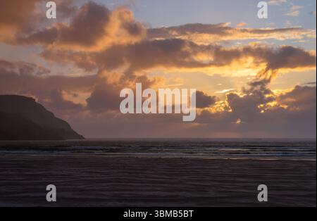 Le coucher du soleil traverse les nuages au-dessus de l'Atlantique à Maghera Beach, le sable ondulé à marée basse et un promontoire silhouette dans le comté de Donegal, en Irlande. Banque D'Images