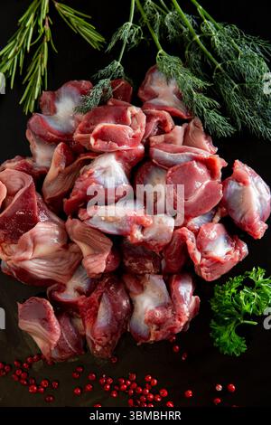Pose à plat de gésiers de poulet crus nettoyés sur une surface noire, décorée de grains de poivre et de légumes verts. Riche en saveur, populaire dans les plats traditionnels. Banque D'Images