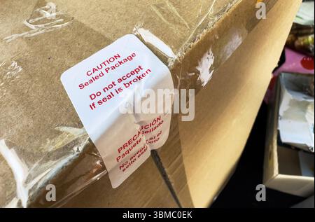 Un gros plan d'un coin d'emballage en carton montre un autocollant d'avertissement blanc avec un texte rouge avertissement de ne pas accepter si le sceau est cassé, sécurisé par un robinet transparent Banque D'Images