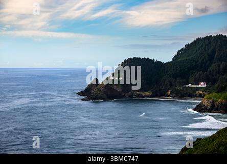 Les vagues s'écrasent contre les falaises rocheuses le long de la côte de l'Oregon, avec un phare historique perché au-dessus des eaux tranquilles et entouré d'une végétation luxuriante Banque D'Images