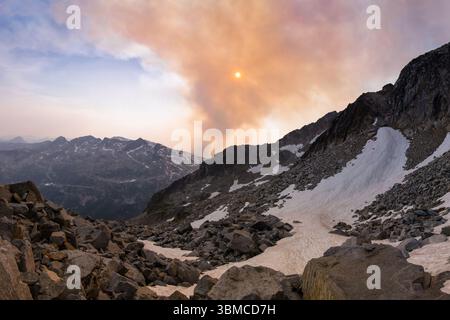 Une vue spectaculaire d'un terrain rocheux où la fumée s'élève d'un feu de forêt dans le parc du glacier de Kokanee, le soleil est partiellement obscurci, projetant une lumière rougeâtre Banque D'Images