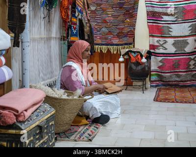 Maroc, Merzouga, 04 juillet 2025. Femme berbère préparant la laine pour le tissage traditionnel de tapis dans un atelier artisanal, mettant en valeur l'artisanat marocain Banque D'Images